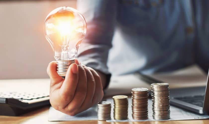 Vīrietis rokās tur spuldzīti blakus monētu kaudzītēm. Vīrietis rokās tur spuldzīti blakus monētu kaudzītēm.